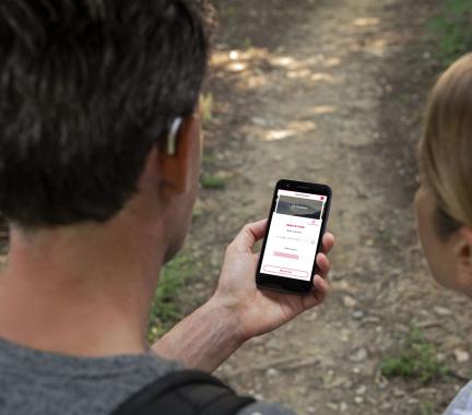 a man and woman on a hiking trail looking at a phone, man is wearing a small, silver hearing aid