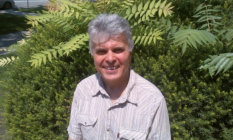 man with gray hair standing in front of a large, green fern outside