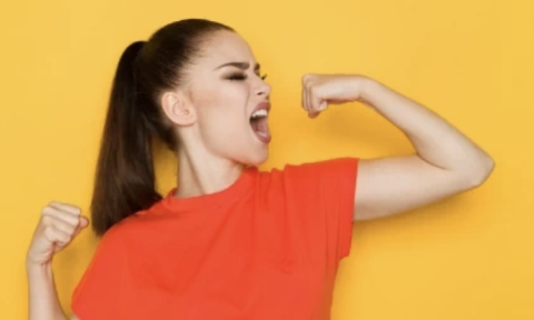 Woman with a brown ponytail flexing her muscles wearing an orange shirt with a yellow background