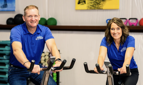 man and woman on stationary bikes looking at the camera and smiling, both are wearing blue shirts