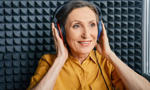 older lady in a sound booth with headphones on, she is touching the ear pieces and wearing a goldenrod shirt