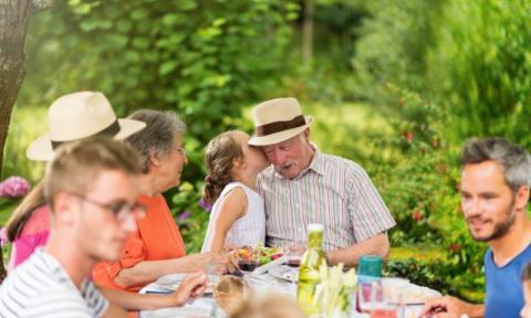 a family at an outdoor picnic with a little girl whispering in her grandpa's ear, he is wearing a wide brim hat with a black hat band