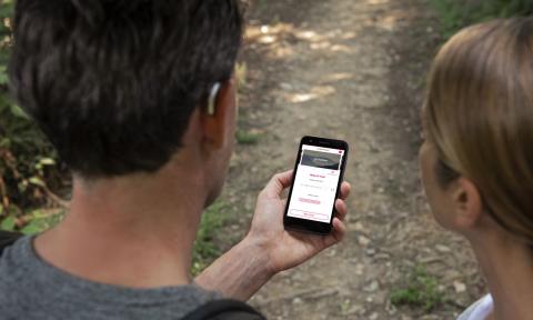 a man and woman on a hiking trail looking at a phone, man is wearing a small, silver hearing aid