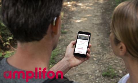 a man and woman on a hiking trail looking at a phone, man is wearing a small, silver hearing aid with amplifon logo in bottom left corner