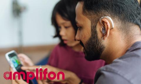 father with hearing aid reading on cell phone screen while daughter sits nearby, with amplifon logo in bottom left corner