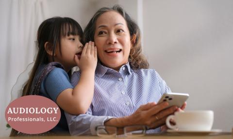 child whispering in elderly woman's ear with logo of audiology professionals llc in bottom left corner