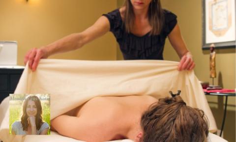 A woman laying on a massage table having her back covered with a sheet, photo of Monica Faux-Kota in bottom left corner