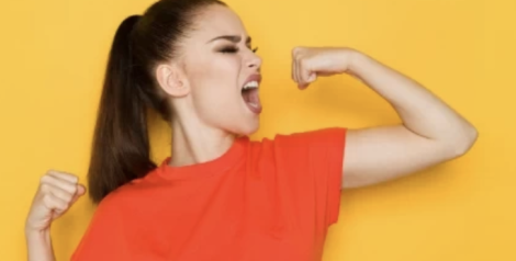 Woman with a brown ponytail flexing her muscles wearing an orange shirt with a yellow background