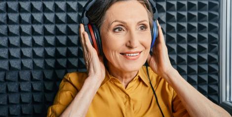older lady in a sound booth with headphones on, she is touching the ear pieces and wearing a goldenrod shirt