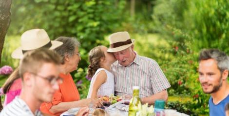 a family at an outdoor picnic with a little girl whispering in her grandpa's ear, he is wearing a wide brim hat with a black hat band