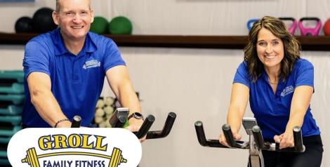 man and woman on stationary bikes looking at the camera and smiling, both are wearing blue shirts, Groll Family Fitness logo bottom left corner