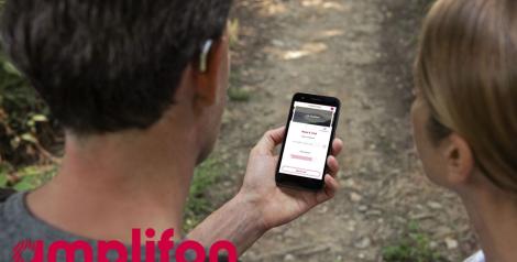 a man and woman on a hiking trail looking at a phone, man is wearing a small, silver hearing aid with amplifon logo in bottom left corner