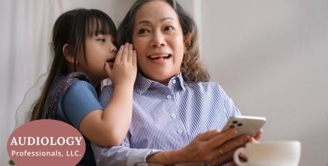 child whispering in elderly woman's ear with logo of audiology professionals llc in bottom left corner