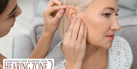 Woman fitting another woman for a hearing aid with Hearing Zone logo in bottom left corner