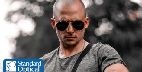 Bald man wearing sunglasses looking down at his watch with Standard Optical logo on bottom left corner