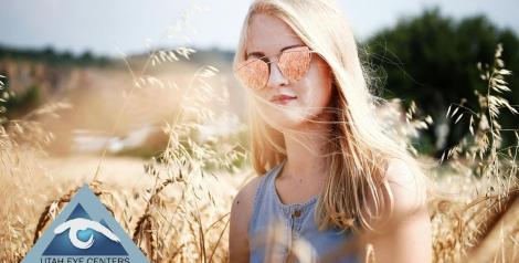 girl with long blonde hair wearing sunglasses in a wheat field with Utah Eye Centers logo in bottom left corner