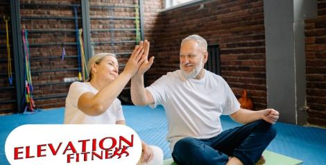 Two older people sitting crosslegged on a mat giving each other a high five, Elevation Fitness logo in bottom left corner