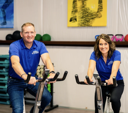 man and woman on stationary bikes looking at the camera and smiling, both are wearing blue shirts